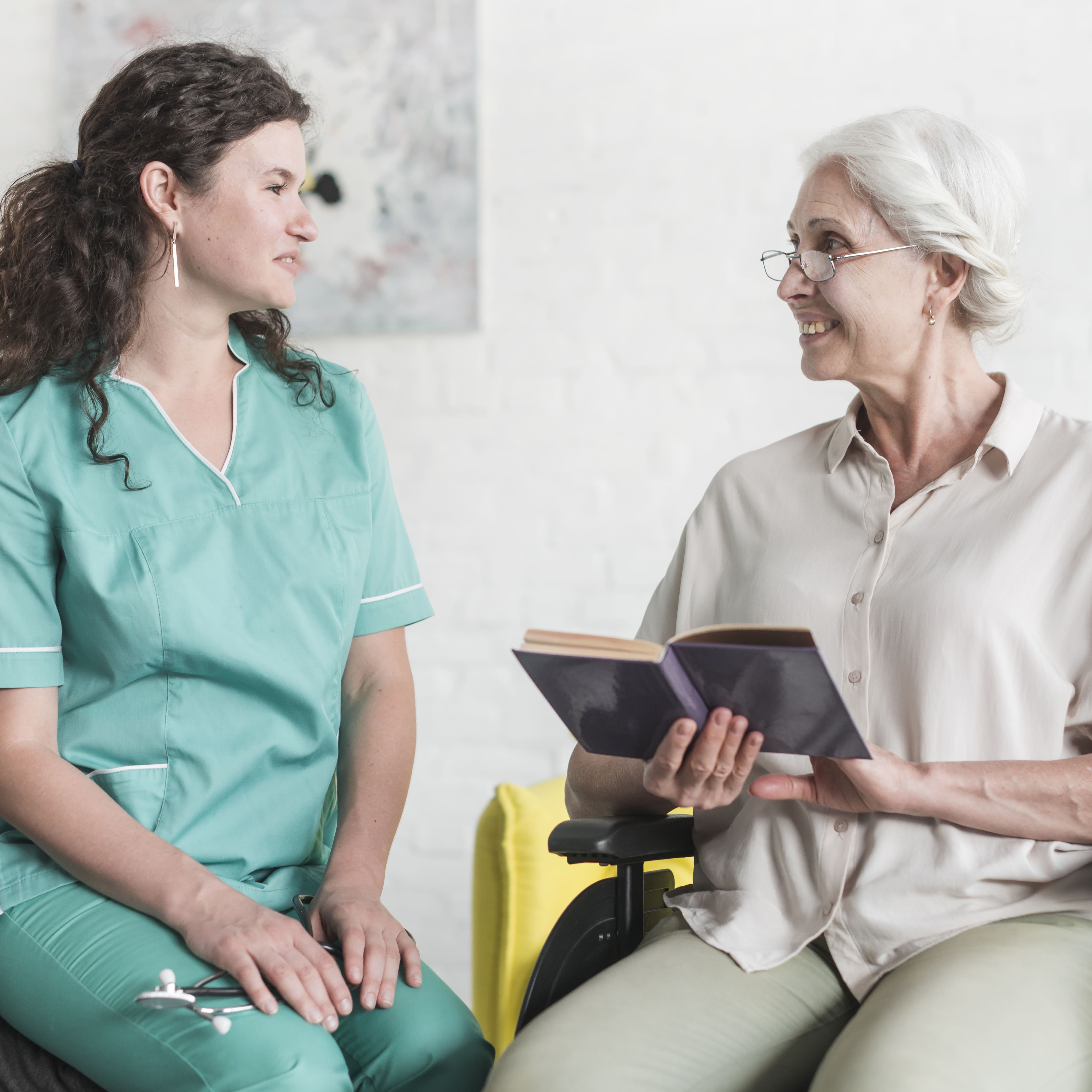 attractive nurse looking senior woman patient sitting wheel chair with book